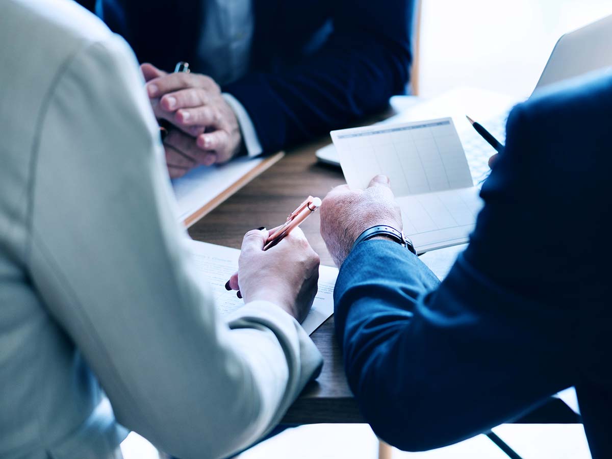 group of business people in suits sitting around a table holding paper and notepads as well as pens discussing and writing strategy