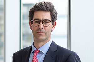 Head shot of Augusta Business man dressed in suit Investment manager in the monitoring team and lawyer Mark de la Haye standing and posing for photo in bright modern London office room next to wide and floor to ceiling windows