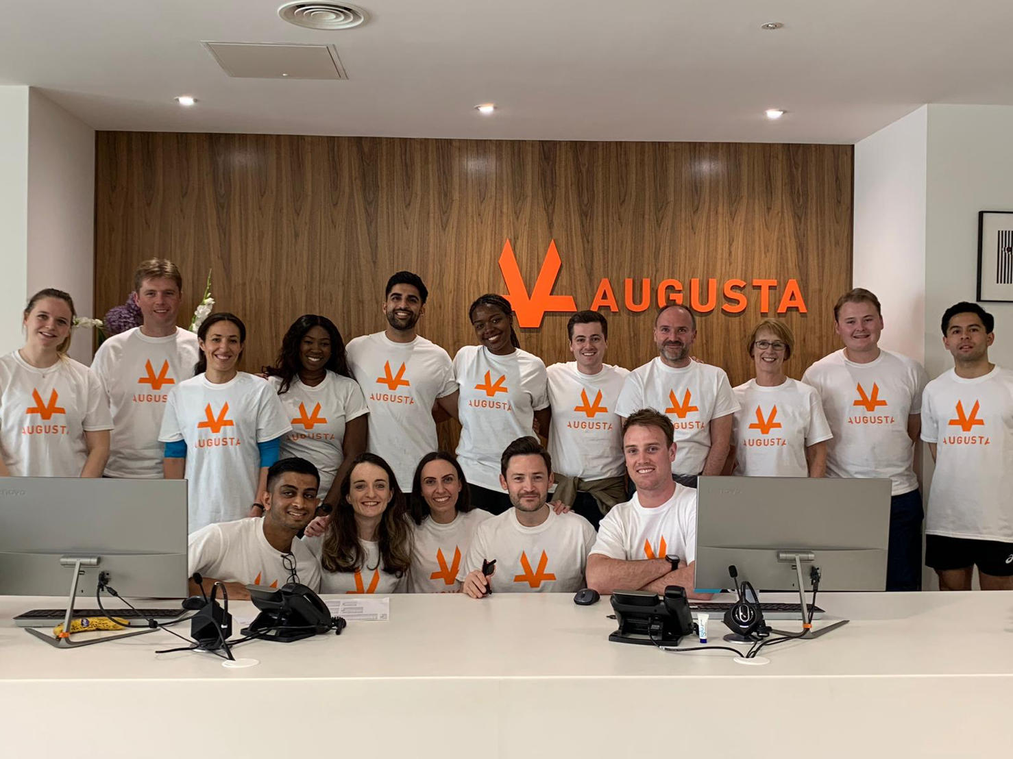 Group photo of 16 members of the Augusta staff lined up in Augusta London office with the orange Augusta Logo in the background smiling and posing for picture with the same Augusta branded T-Shirt