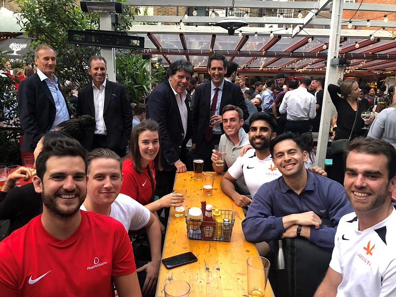 Group photo taken outside around a pub table in daylight with people sat around and standing smiling at camera with drinks