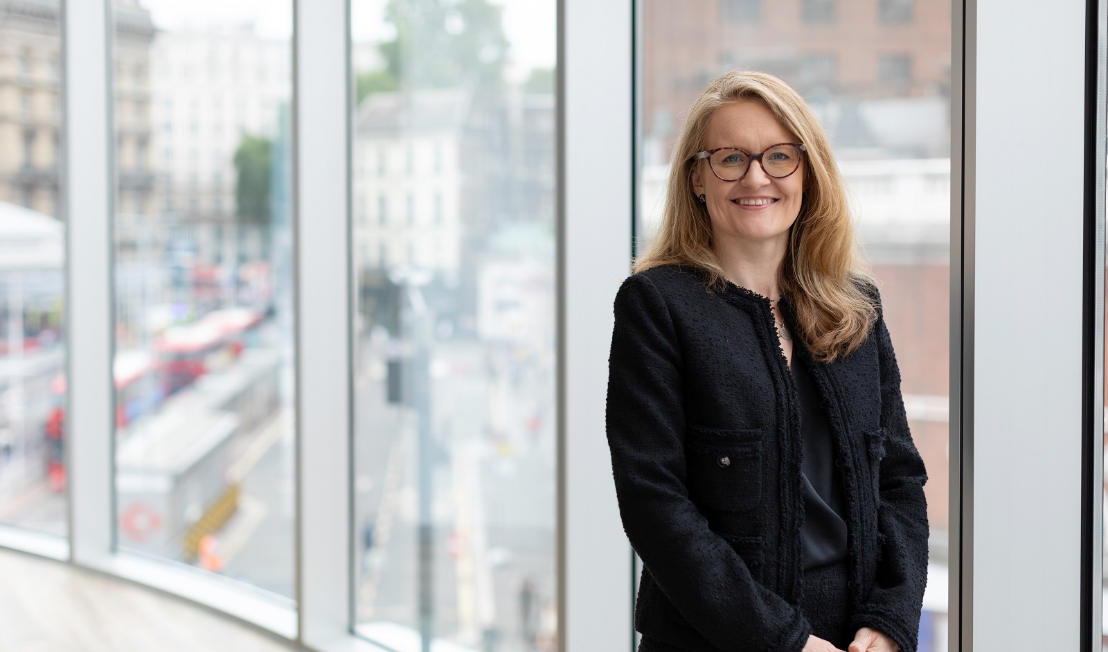 Augusta Business women dressed in formal dress Chief Operating Officer COO Polly Bahl standing and posing for photo with a smile in bright modern London office room next to wide and floor to ceiling windows