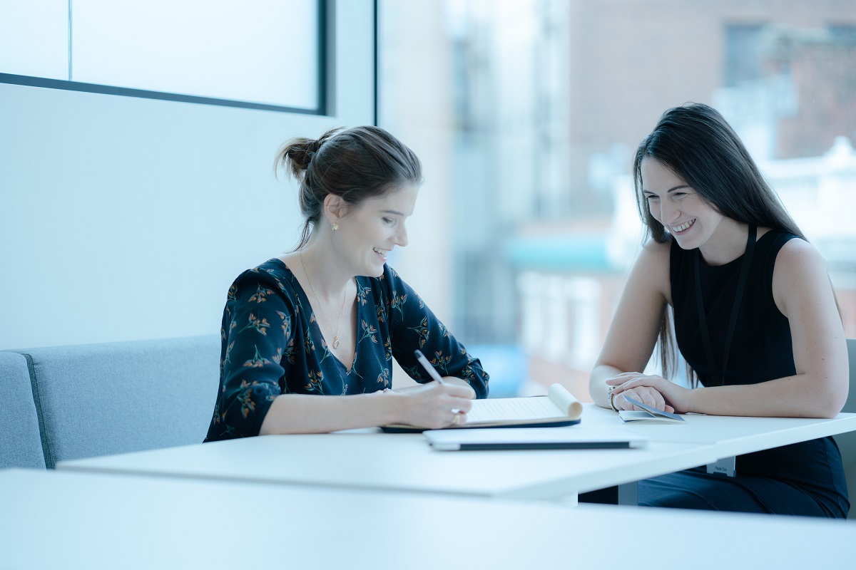 Business women and investment managers at Augusta sitting at table speaking and laughing over documents with pens to hand in front of a bright floor to ceiling window with buildings outside